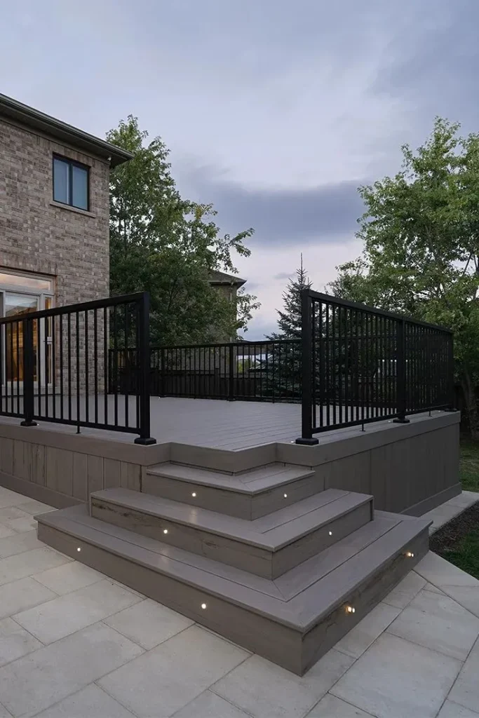 Multilevel gray composite deck with black railings and recessed lighting on the stairs, next to a brick house.